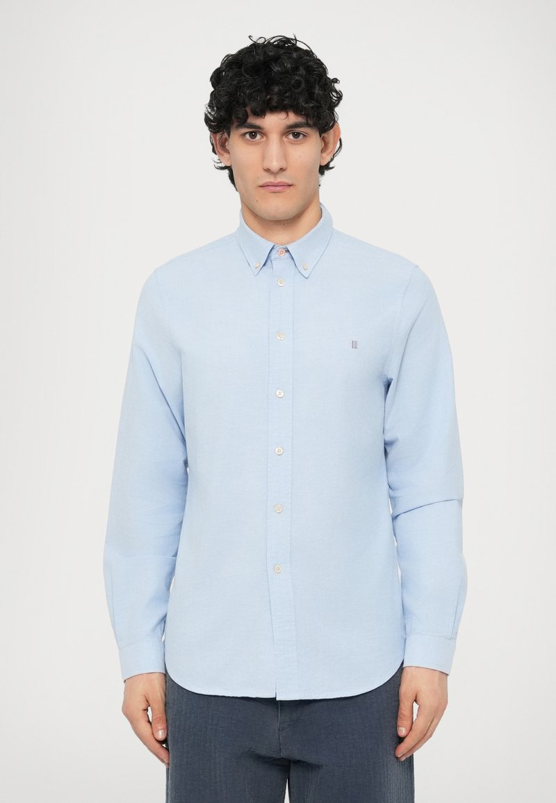 Vaaleansininen pitkähihainen nappipaita, valmistettu teksturoidusta kankaasta. Päällä pieni logo, pyöristetty helma ja kontrastiväriset napit.