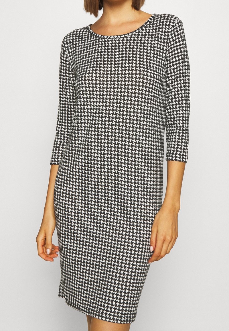 Femme portant une robe longueur genou à motif pied-de-poule noir et blanc, manches trois quarts et encolure ronde.