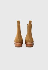 Botas de tobillo de ante color beige con un tacón bajo, que presentan paneles laterales elásticos y una suela de madera lisa, con detalles de costura visibles.