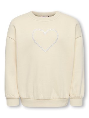 Jeune fille aux longs cheveux portant un sweat-shirt beige avec un motif en forme de cœur, pantalon gris et baskets beiges, debout les mains dans le dos.