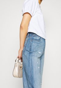 Blusa blanca corta con mangas abullonadas combinada con jeans desgastados de un azul claro. Sosteniendo un pequeño bolso beige con herrajes plateados.