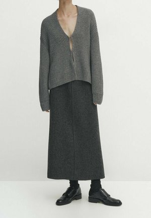 Femme portant un cardigan côtelé gris, une longue jupe gris foncé, des chaussettes noires et des chaussures en cuir noires, debout devant un fond blanc uni.