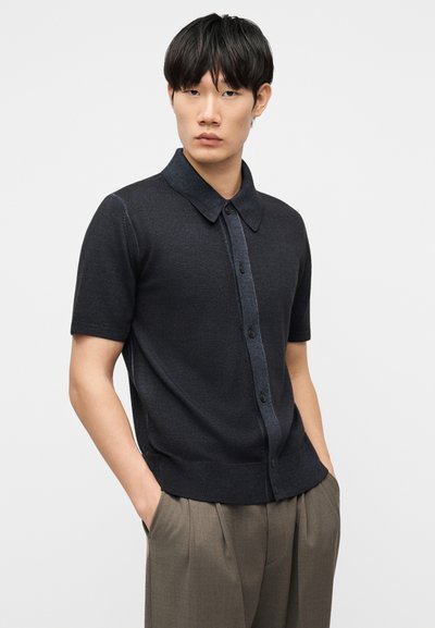 Νεαρός άνδρας με μαύρα μαλλιά, ντυμένος με σκούρο κοντομάνικο πουκάμισο με κουμπιά και καφέ πιετέ παντελόνι, τα χέρια στις τσέπες, ουδέτερο φόντο.