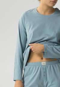 Jasnoniebieska bluzka z długim rękawem, okrągłym dekoltem i miękkiego materiału, w połączeniu z pasującymi jasnoniebieskimi spodniami z elastyczną talią i guzikami.