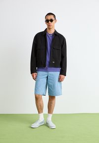 Sort zip-up jakke med to frontlommer, lilla t-shirt under, lyseblå denimshorts og grå ternede slip-on sko.