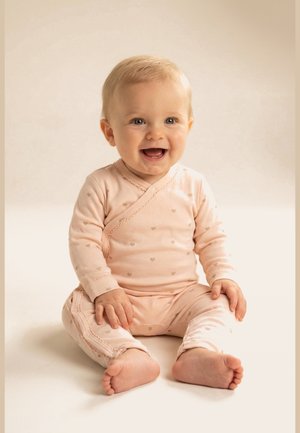 Uśmiechnięte niemowlę z jasnymi włosami, siedzące, ubrane w różowy strój z długim rękawem i małe wzory serc na neutralnym tle.