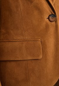 Veste en daim marron en gros plan montrant une poche à rabat et un bouton marron foncé à quatre trous près du revers.