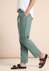 Helle grüne Hosen mit einem lockeren Schnitt, ausgestattet mit Seitentaschen und einem sichtbaren Bund. Dazu schwarze Sandalen mit Leopardenmuster und Schnallen.