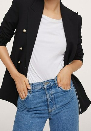 Person trägt einen schwarzen Blazer mit goldenen Knöpfen, ein weißes T-Shirt und blaue Jeans mit den Händen in den vorderen Taschen.