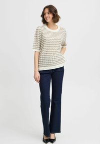 Suéter de punto con mangas cortas, color crema, presenta un patrón ondulado con acentos beige. Combinado con pantalones de mezclilla oscuros de pierna acampanada y tacones negros.