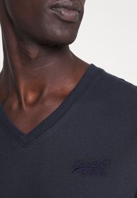 Námořnická modrá bavlněná trička s výstřihem do V a žebrovaným límcem. Obsahuje fialové vyšívané logo "Superdry" na hrudi. Hladká textilie, přiléhavý design.