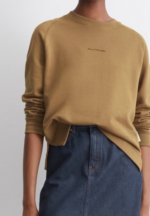 Personne portant un sweat-shirt à manches longues couleur beige avec un petit texte central, assorti à un pantalon en denim bleu foncé taille haute.