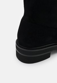 Schwarze Wildleder-Stiefelette mit strukturiertem Finish, die über eine dicke schwarze Sohle und ein dezentes horizontales Streifendetail an der Ferse verfügt.