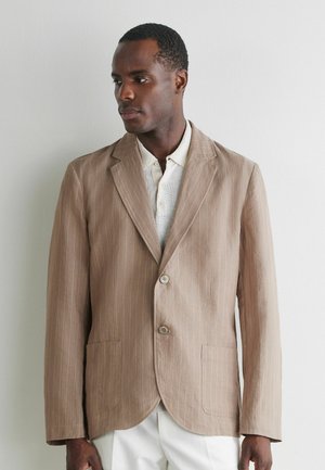 Mann trägt beige Nadelstreifenjacke und weißes Strick-Poloshirt, steht vor einer schlichten hellen Wand, schaut nach links.