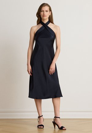 Femme portant une robe noire sans manches à col halter croisé sur le devant et des sandales noires à talons hauts, debout sur un sol en bois clair.