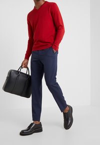 Mężczyzna w czerwonym swetrze w serek, granatowych spodniach i czarnych eleganckich butach, trzymający czarną skórzaną teczkę, stojący na jednolitym tle.