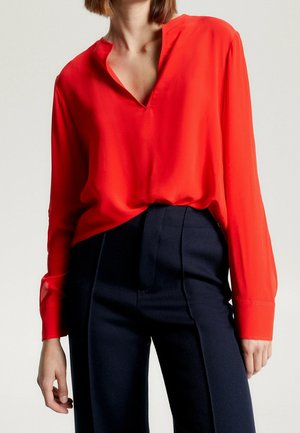 Blusa rossa a maniche lunghe con scollo a V, realizzata in un tessuto morbido; abbinata a pantaloni strutturati a vita alta di un blu navy scuro.