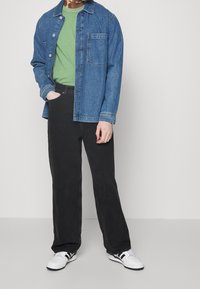 Džínová bunda středně modré barvy přes zelené tričko, spárovaná s černými širokými kalhotami a bílými teniskami se černými pruhy.