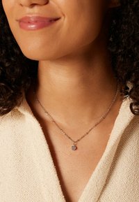 Zilveren ketting met een cirkelvormige hanger met een sprankelend detail, gedragen met een lichtgekleurde textuurstof.