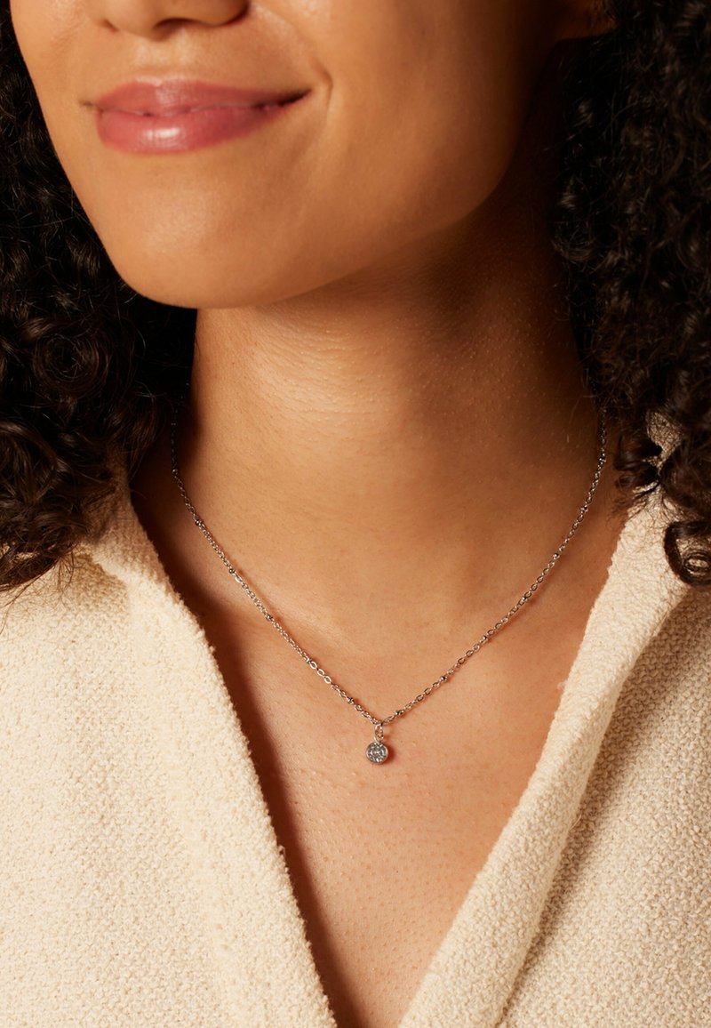 Zilveren ketting met een cirkelvormige hanger met een sprankelend detail, gedragen met een lichtgekleurde textuurstof.