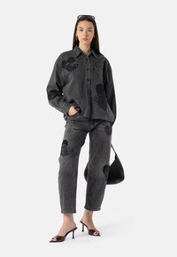 Čierna denimová košeľa a nohavice s veľkými textúrovanými čiernymi aplikáciami v tvare srdca. Modelka má na sebe sandále s otvorenou špičkou a drží čiernu kabelku.