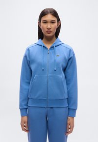 Φως μπλε φούτερ με φερμουάρ, κατασκευασμένο από μαλακό ύφασμα, με τσέπη καγκουρό, ριμπ μανσέτες και μικρό κεντημένο λογότυπο στο αριστερό στήθος.