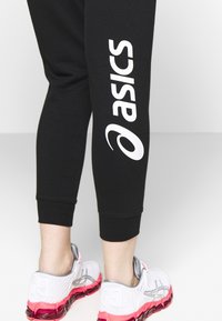 Zwarte sportleggingen met een witte "ASICS" logo aan de zijkant, gecombineerd met wit en roze hardloopschoenen met gestructureerde accenten.