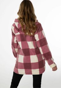 Giacca in pile a quadri con grandi quadrati rosa e crema, cappuccio, maniche lunghe e vestibilità comoda. Sotto si indossano pantaloni neri.