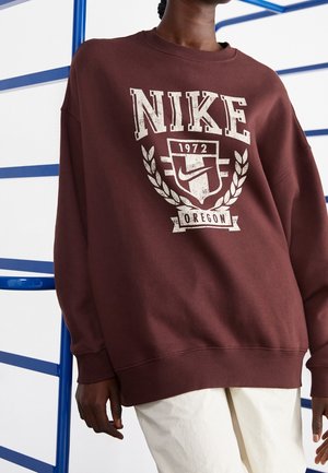 Persoon die een kastanjebruwe Nike-trui draagt met de tekst "1972 Oregon" en logo, staand bij een blauwe ladderachtige constructie.