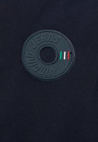 Svart tyg med en texturerad yta som har en rund präglad gummilogga som säger "JEANS" och inkluderar ett hål i mitten.