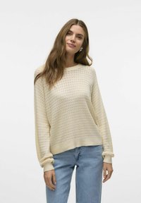 Maglione a maglia color crema con un motivo testurizzato, maniche ampie e polsini a coste, abbinato a jeans in denim azzurro chiaro.