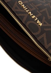 Braune gemusterte Reißverschlusstasche mit einem Logo und goldenen Beschlägen, mehreren Fächern, präsentiert eine glatte Textur und ein sauberes Design.