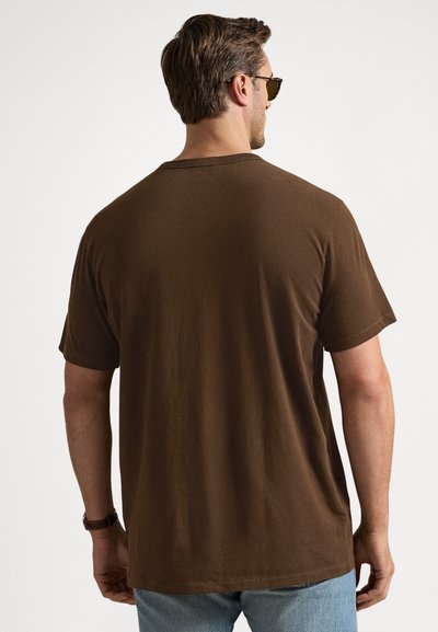 Καφέ βαμβακερό t-shirt με στρογγυλή λαιμόκοψη και κοντά μανίκια, με άνετη εφαρμογή. Φορεμένο με ανοιχτόχρωμο μπλε τζιν, από πίσω όψη.