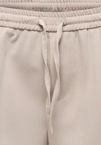 Pantalon en tissu beige avec taille élastique cousue et long cordon non noué suspendu au centre devant.
