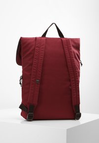 Mochila de tela roja con diseño de tapa enrollable, correas negras y un asa. Cuenta con correas de hombro acolchadas ajustables y un broche lateral.