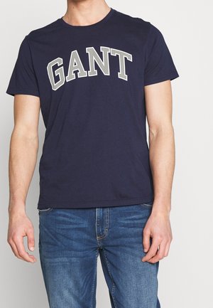 Laivastonsininen puuvillainen t-paita, jossa suuri kaareva "GANT"-logo harmaana rinnan yli. Lyhyet hihat ja pyöreä pääntie.