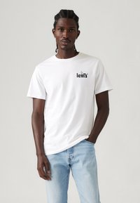 Weißes Baumwoll-T-Shirt mit rundem Halsausschnitt und kurzen Ärmeln, versehen mit einem schwarzen Drucklogo auf der Brust, kombiniert mit hellblauen Jeans.