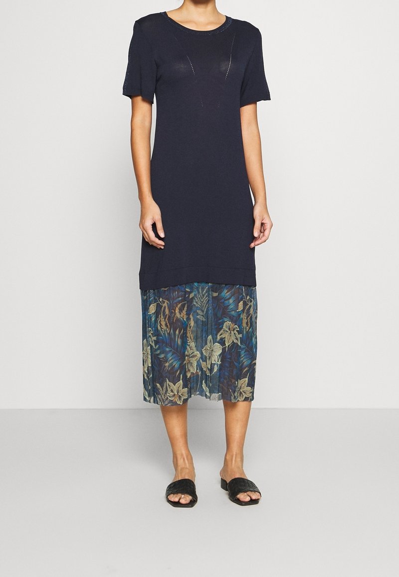 Femme portant une robe courte bleu marine à manches courtes avec une jupe midi transparente à motif floral et des sandales noires tissées sans lacets, debout devant un fond uni.