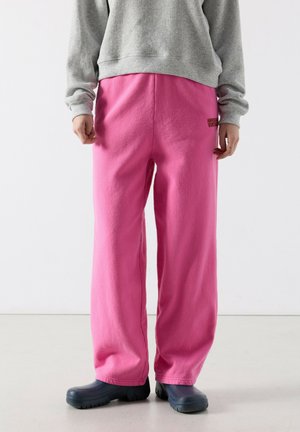 Apatinė kūno dalis dėvinčio asmens, kuris vilki laisvas ryškiai rožines laisvalaikio kelnes su juodu užrašu dešinėje šlaunies pusėje, pilką džemperį ir juodus guminį slip-on tipo batus.