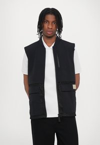Black sleeveless vest with zip closure, textured side panels, and two front pockets. Worn over a white polo shirt.