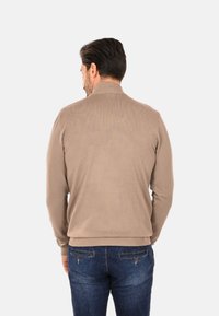 Maglione beige lavorato a maglia con collo alto, texture a coste e design aderente, abbinato a jeans blu scuro. Vista posteriore.