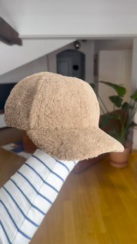 Șapcă maro, texturată, cu o coroană rotunjită și o vizieră scurtă, curbată. Materialul pare moale și pufos. Ținută în mână pe un fundal deschis la culoare.