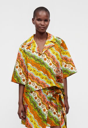 Personne portant une chemise et une jupe assorties colorées avec des motifs floraux et tourbillonnants orange, vert, jaune et blanc, se tenant devant un fond uni.