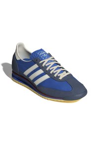 Blue athletic shoe with grey accents, three white stripes, textured blue tongue, and a rubber sole with yellow tread.