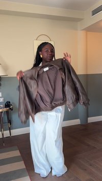 Brun läderjacka med ribbad elastisk nederkant, uppvisad öppen för att visa ett slätt innerfoder. Vita vida byxor fulländar outfiten.