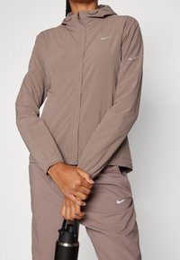 Chaqueta Nike ligera en taupe con capucha, cierre de cremallera y puños elásticos. Presenta detalles reflectantes sutiles y un tejido suave y liso.