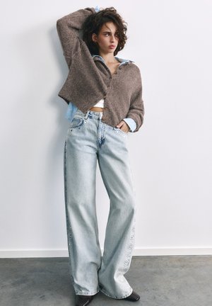 Jeune femme aux cheveux bouclés porte un cardigan marron boutonné sur une chemise en denim et un jean large délavé clair avec broderies florales, debout contre un mur blanc.