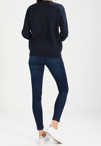 Pull en maille bleu marine foncé, avec un col rond et des épaules tombantes. Jean bleu foncé ajusté, se terminant à la cheville, associé à des baskets blanches.