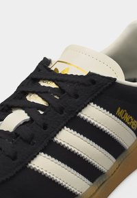 Κοντινό πλάνο σε μαύρο και άσπρο αθλητικό παπούτσι Adidas με μαύρα κορδόνια, άσπρες ρίγες, χρυσό κείμενο "MÜNCHEN" και καφέ σόλα.