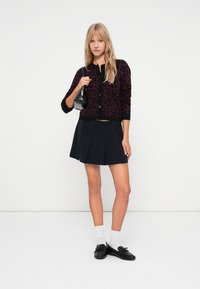Dark purple leopard-print cardigan with button closure, paired with a navy pleated mini skirt. Model holds a small black handbag.